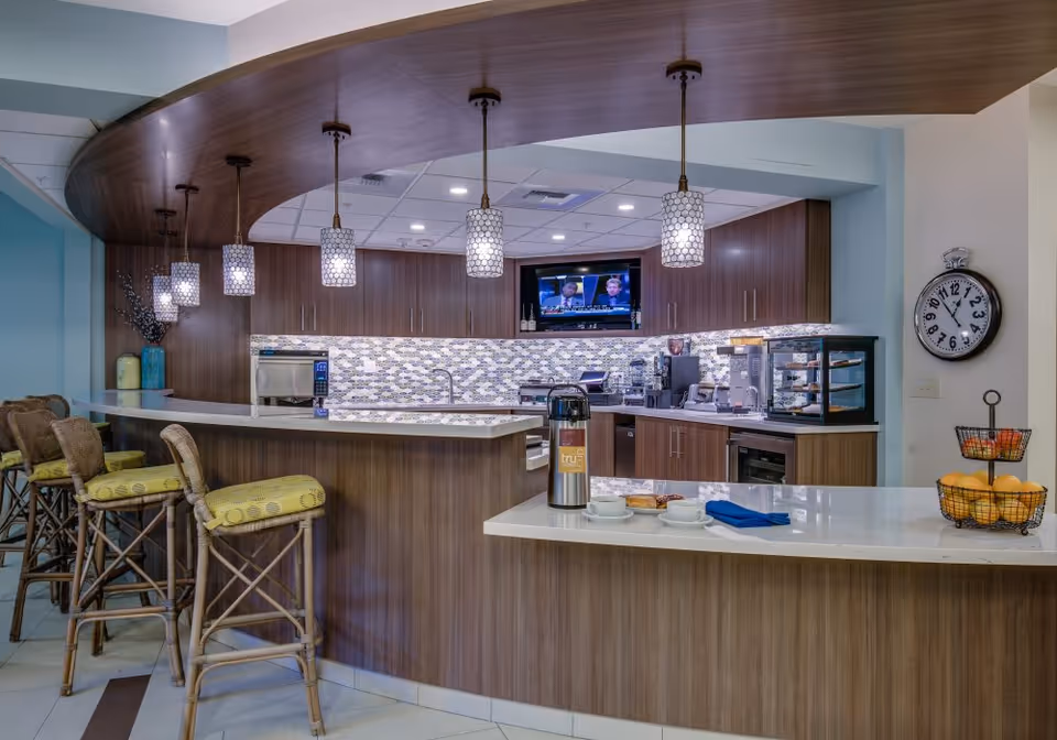 A modern kitchen area in a senior living facility with a curved wooden counter and four bar stools with yellow cushions. The kitchen features a mosaic tile backsplash, multiple pendant lights hanging from a wooden ceiling accent, a wall-mounted TV, and various kitchen appliances. On the counter, there is a coffee dispenser, cups, plates, and a two-tiered fruit basket with oranges and apples. A large round clock is mounted on the wall to the right.