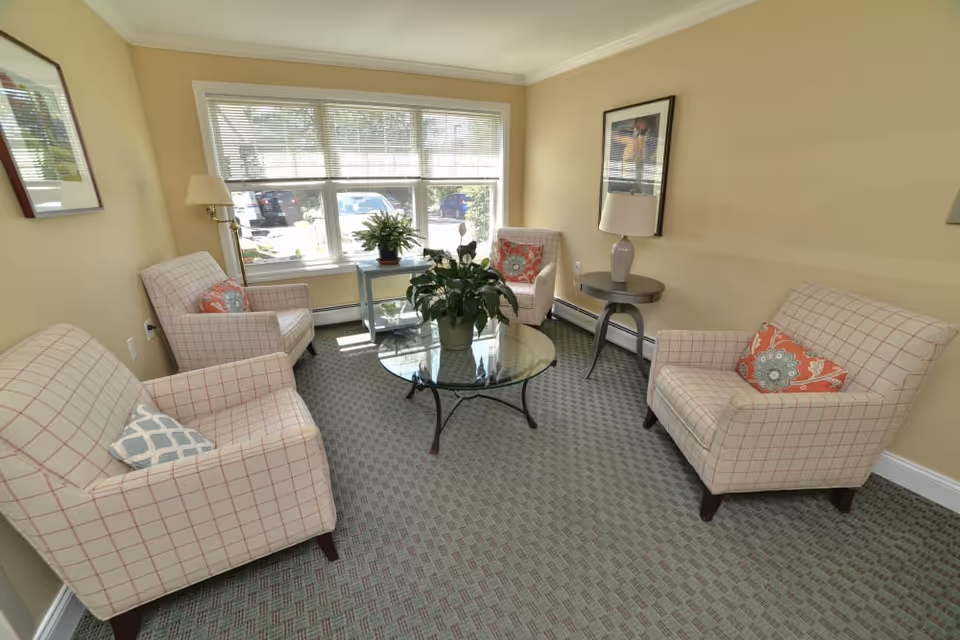 A cozy living room with four beige armchairs featuring a red grid pattern, each with decorative pillows. A round glass coffee table with a potted plant sits in the center on a patterned carpet. There is a large window with blinds letting in natural light, a small side table with a lamp, and framed artwork on the walls.