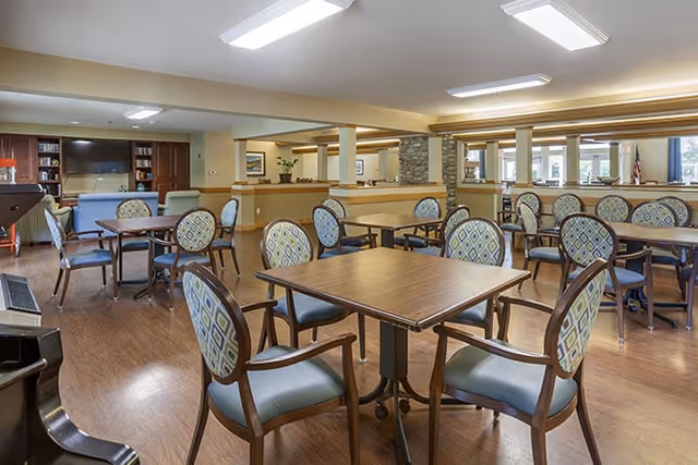 A spacious dining room with multiple wooden tables and cushioned chairs arranged neatly. The room has wood flooring, beige walls, and ceiling lights. In the background, there is a seating area with a blue couch and a large flat-screen TV mounted on a wooden cabinet. Large windows allow natural light to enter the room.