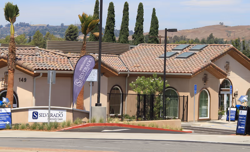 Exterior view of Silverado Brea Memory Care Community building with beige walls and a tiled roof, surrounded by palm trees and other greenery. There are signs and banners displaying the facility's name near the entrance, and a parking area with handicap parking spaces is visible.