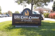 Outdoor view of a sign for Life Care Center of Greeley, a skilled nursing and Alzheimer's care facility, situated on a grassy area with trees, bushes, and a building in the background.