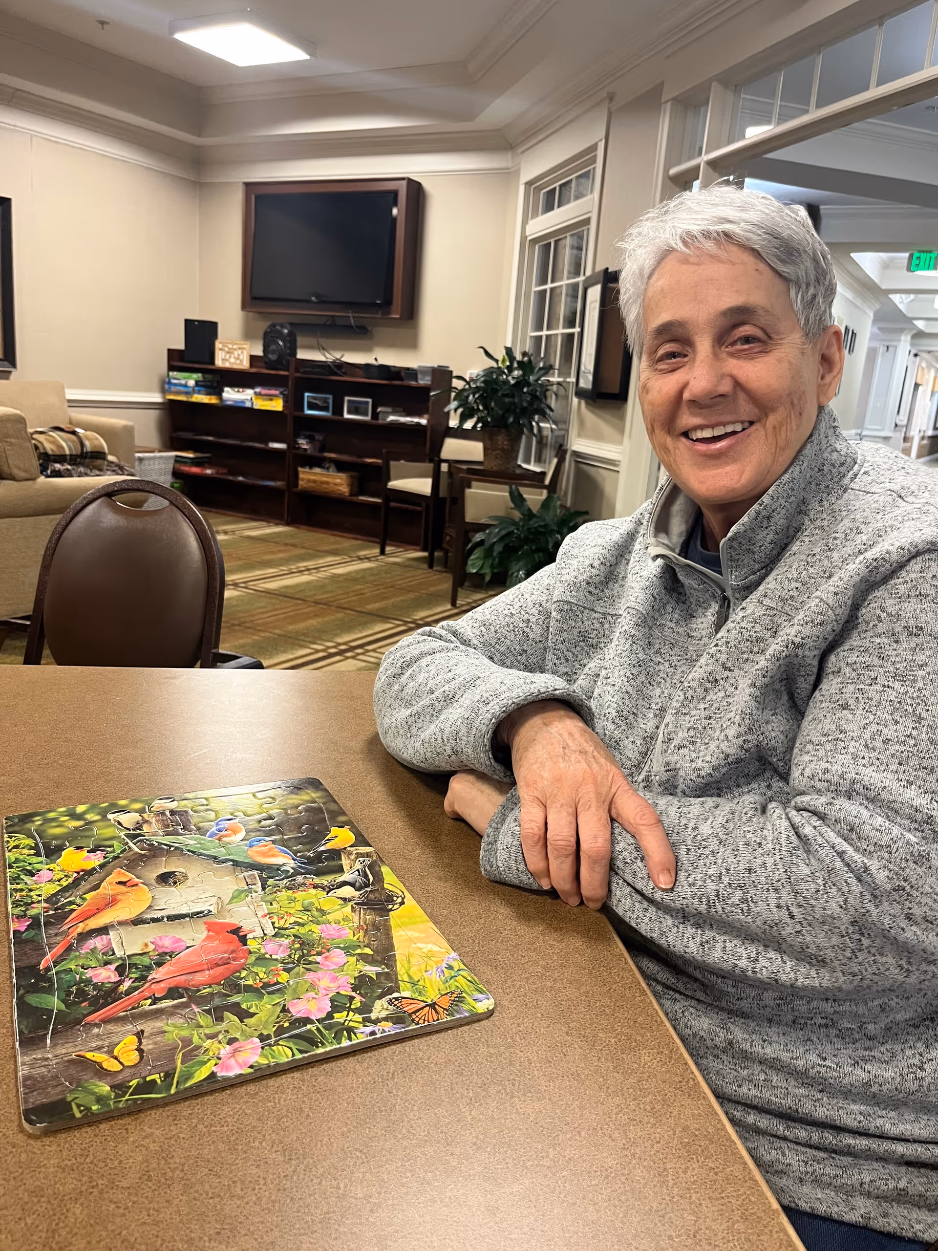 An elderly person with short gray hair and a gray sweater sits at a table smiling. On the table is a colorful completed jigsaw puzzle featuring birds, flowers, and butterflies. The background shows a cozy room with a TV mounted on the wall, bookshelves, chairs, plants, and a hallway.