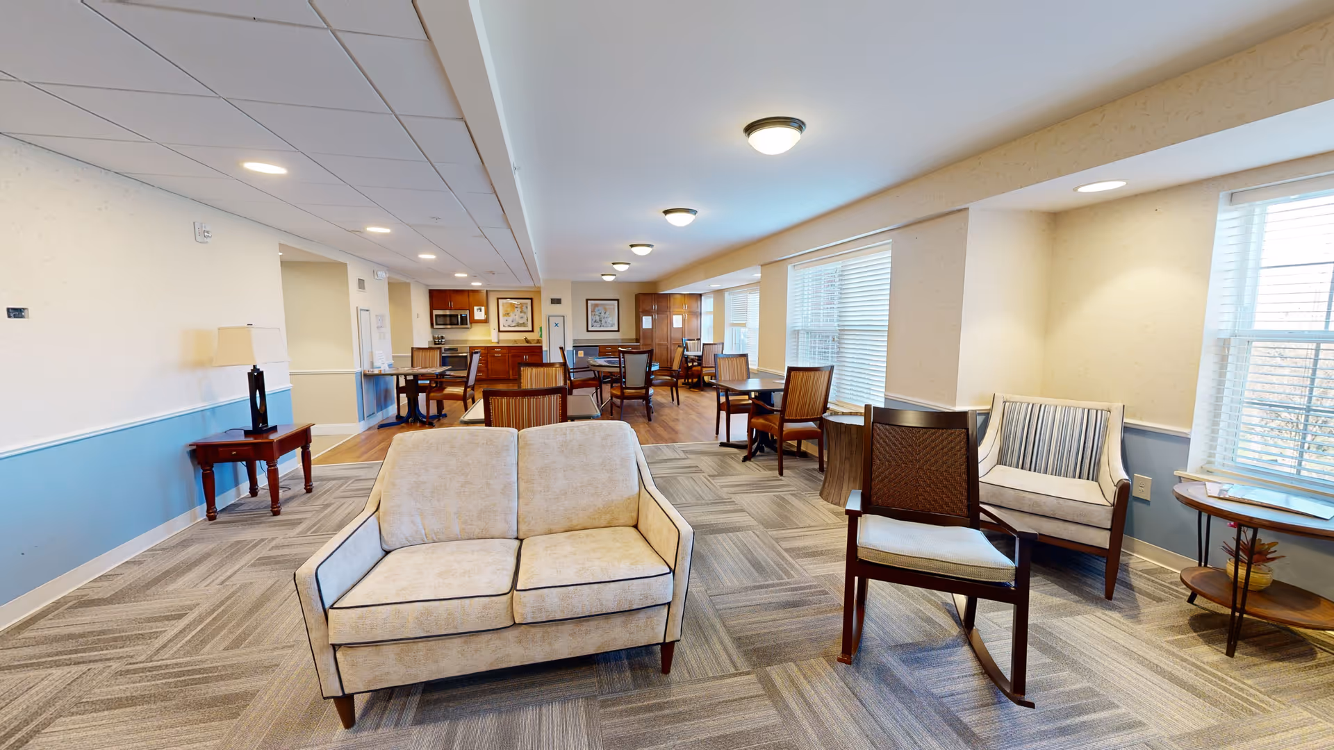 A spacious senior living common area with beige and blue walls, carpeted and wooden flooring, several chairs and tables, a small sofa, and a kitchenette with cabinets, microwave, and artwork on the walls.