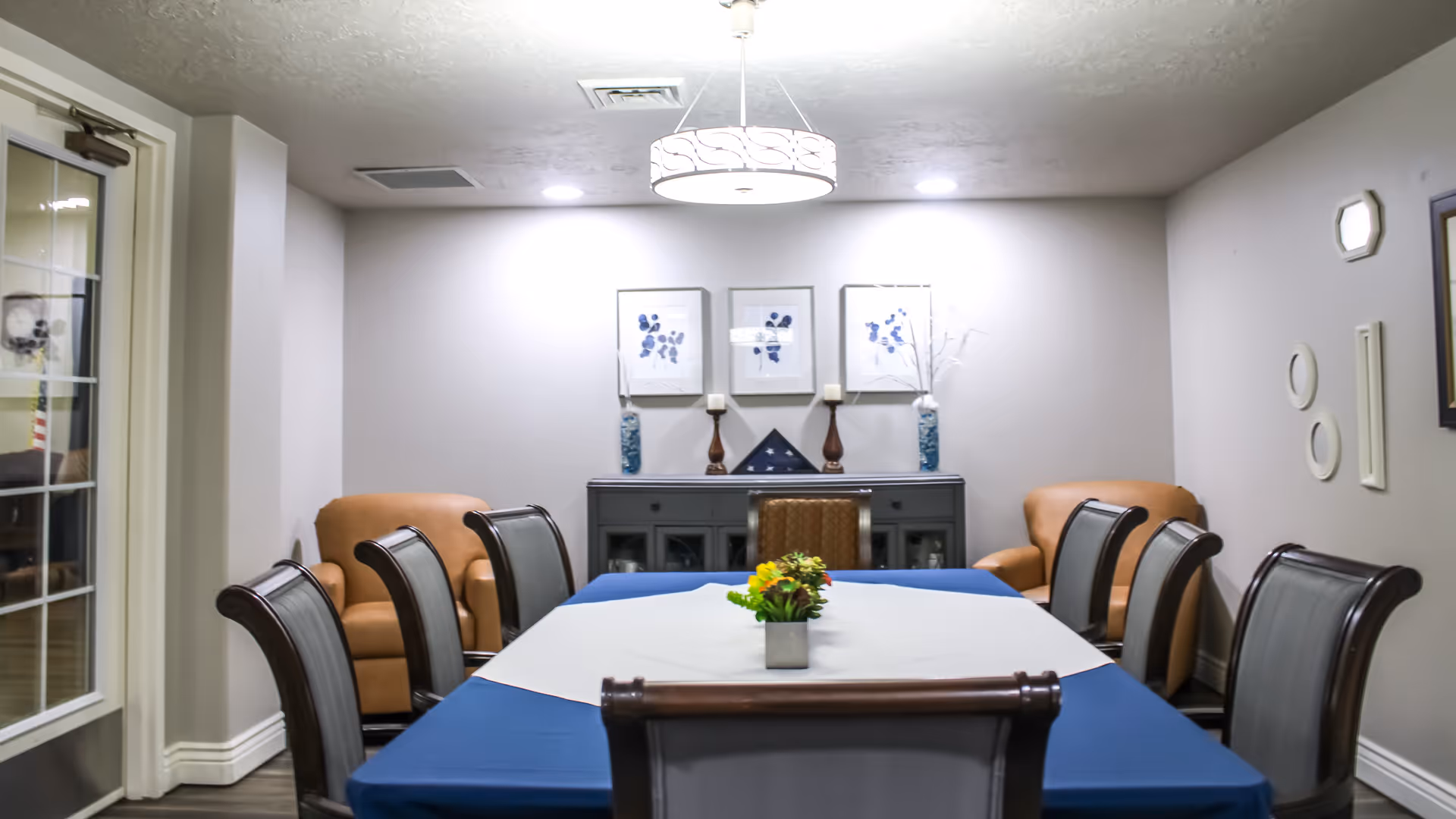 A dining room with a rectangular table covered with a blue tablecloth and a white runner, surrounded by eight chairs. Two tan armchairs are placed against the back wall, which also features a dark sideboard with decorative items including two blue vases, two candles, and a folded American flag. Three framed floral artworks hang above the sideboard. The room is lit by a ceiling light fixture and recessed lights.