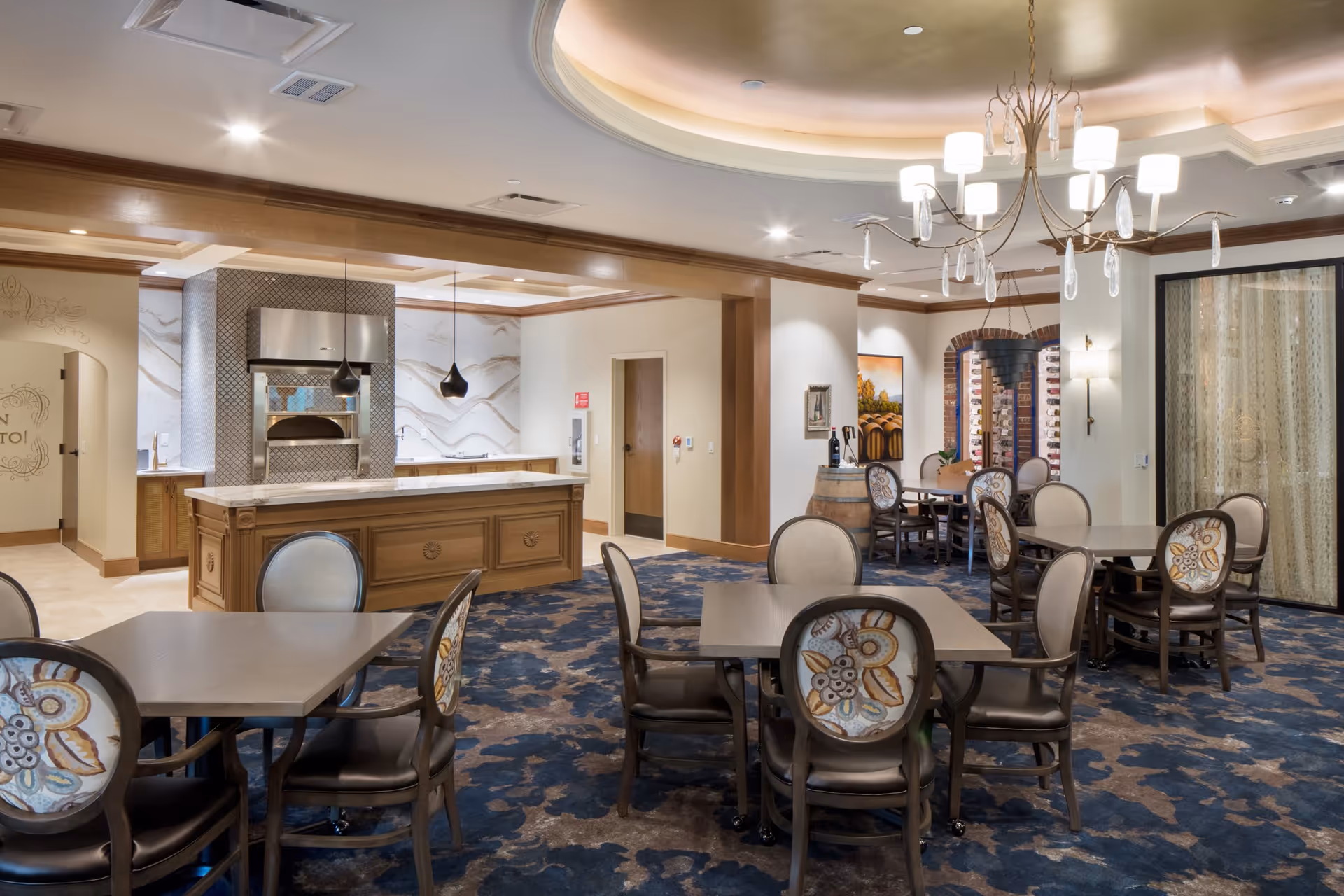 A spacious and elegant dining area with multiple tables and chairs featuring floral upholstery. The room has a decorative ceiling with a chandelier, a kitchen area with a marble countertop and modern appliances, and a wine display in the background.