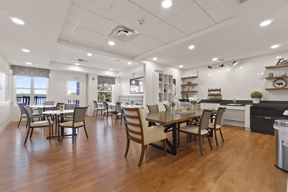 A bright and spacious dining area with wooden floors, several tables and chairs, large windows with views of greenery and water outside, and a coffee station with white subway tile backsplash and open shelves.
