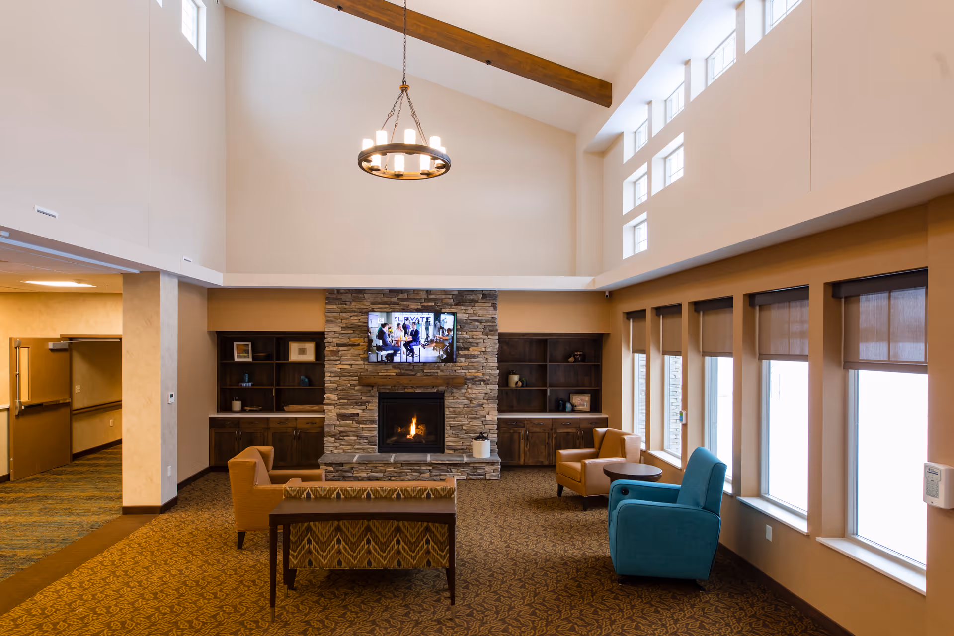 Spacious senior living common lounge with high ceilings, a stone fireplace and TV, multiple chairs and sofas, and large windows.