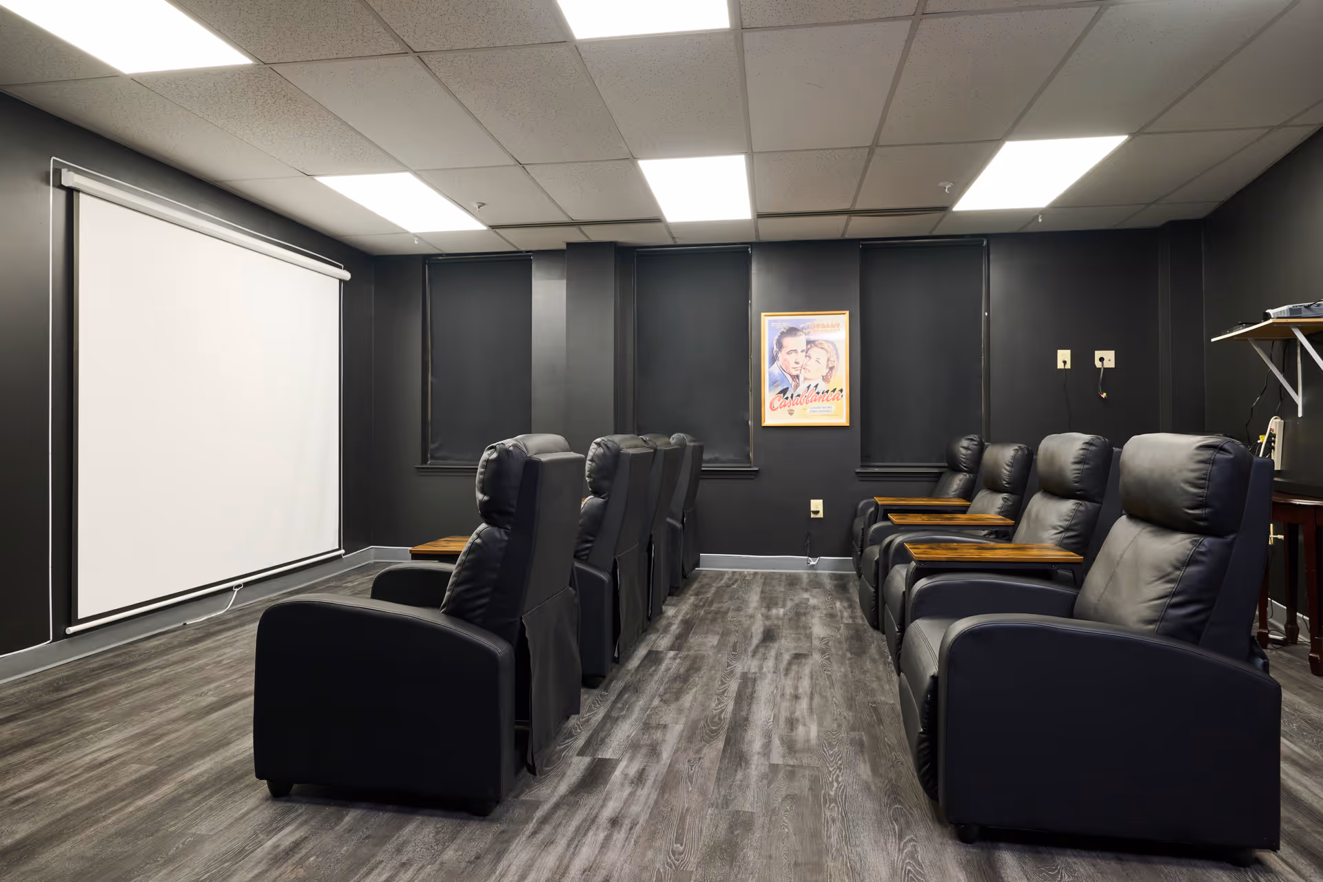 A small movie theater room with two rows of black leather recliner chairs facing a large white projection screen. The walls are painted black with three windows covered by black blinds. A framed Casablanca movie poster is hung on the wall between the windows. The floor has dark wood-style flooring and the ceiling has recessed lighting panels.