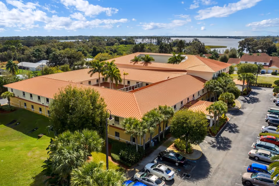 Aerial view of a two-story Mediterranean-style assisted living building with red tile roofs, palm trees, parked cars, and water in the background.