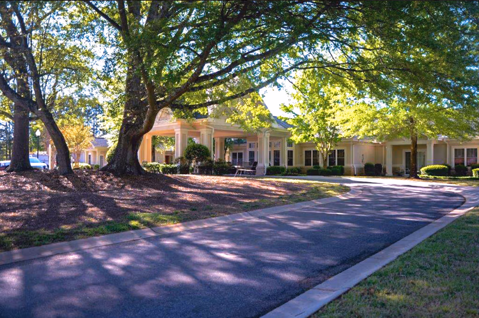 A curved driveway leads to the entrance of a single-story building surrounded by large trees with green foliage casting shadows on the ground. The building has a covered porch area with columns and several windows, set in a landscaped area with bushes and grass.