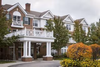 Exterior view of a multi-story senior living facility building with white balconies, large windows, and a covered entrance. The landscaping includes green trees and bushes with some autumn-colored foliage in the foreground.