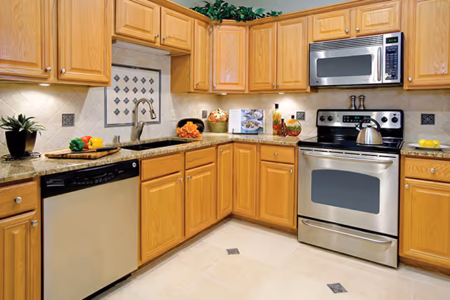 A modern kitchen with wooden cabinets, granite countertops, a stainless steel dishwasher, oven, and microwave. The kitchen features a sink with a faucet, decorative tiles on the backsplash, and various kitchen items including a kettle, vegetables, and a cookbook on the counter.