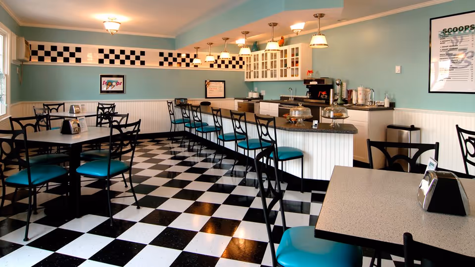 Bright retro-style dining area with black-and-white checkered floor, teal-upholstered chairs, tables and a counter with pendant lights.