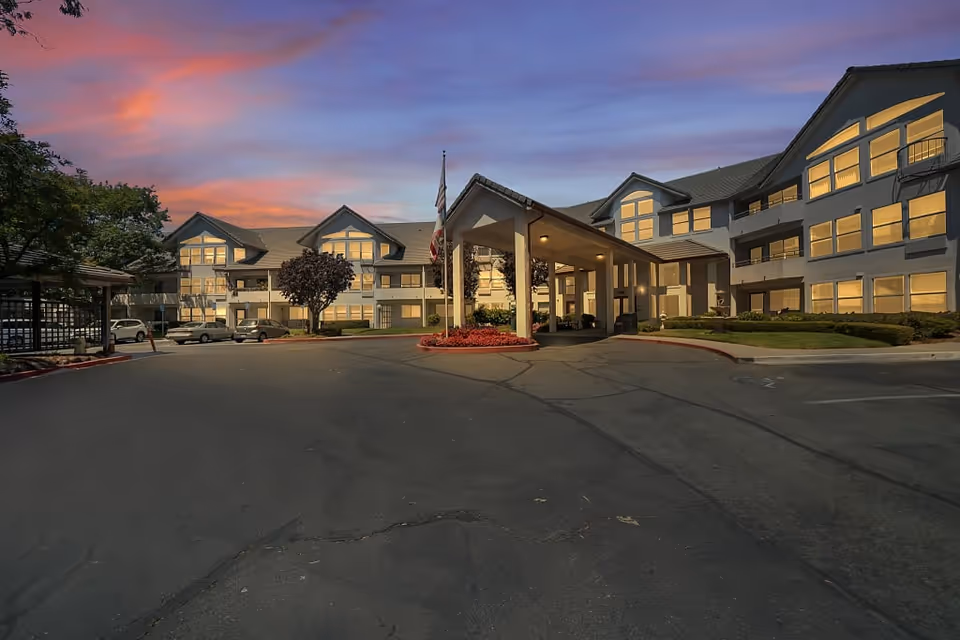 Exterior view of Truewood by Merrill, Pinole senior living facility at sunset with a driveway, entrance canopy, parked cars, and well-lit windows.