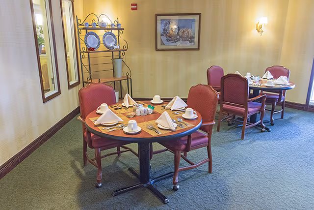 A dining area with two round tables set for four people each, featuring red cushioned chairs, white napkins folded on plates, cups, and silverware. The room has light yellow striped wallpaper, a framed picture on the wall, and a decorative plate rack with plates displayed.