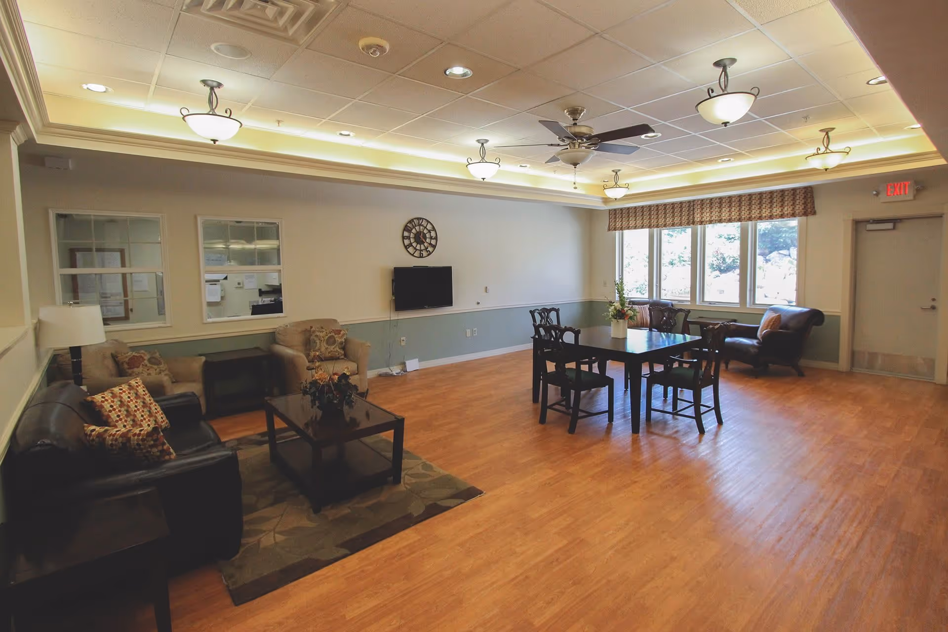 A spacious living room area with wooden flooring, featuring a black leather sofa with patterned pillows, two beige armchairs, a coffee table with a floral arrangement, a dining table with four chairs, a wall-mounted TV, a large clock, ceiling fans, and multiple ceiling lights. Large windows with patterned valances allow natural light to fill the room.
