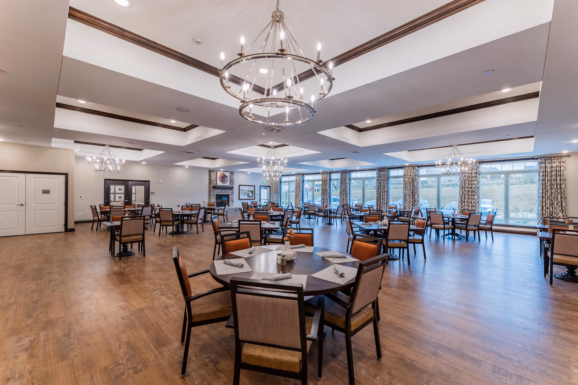 Spacious dining room with multiple round and square tables surrounded by chairs, large windows with patterned curtains allowing natural light, wooden flooring, and modern chandeliers hanging from a coffered ceiling.