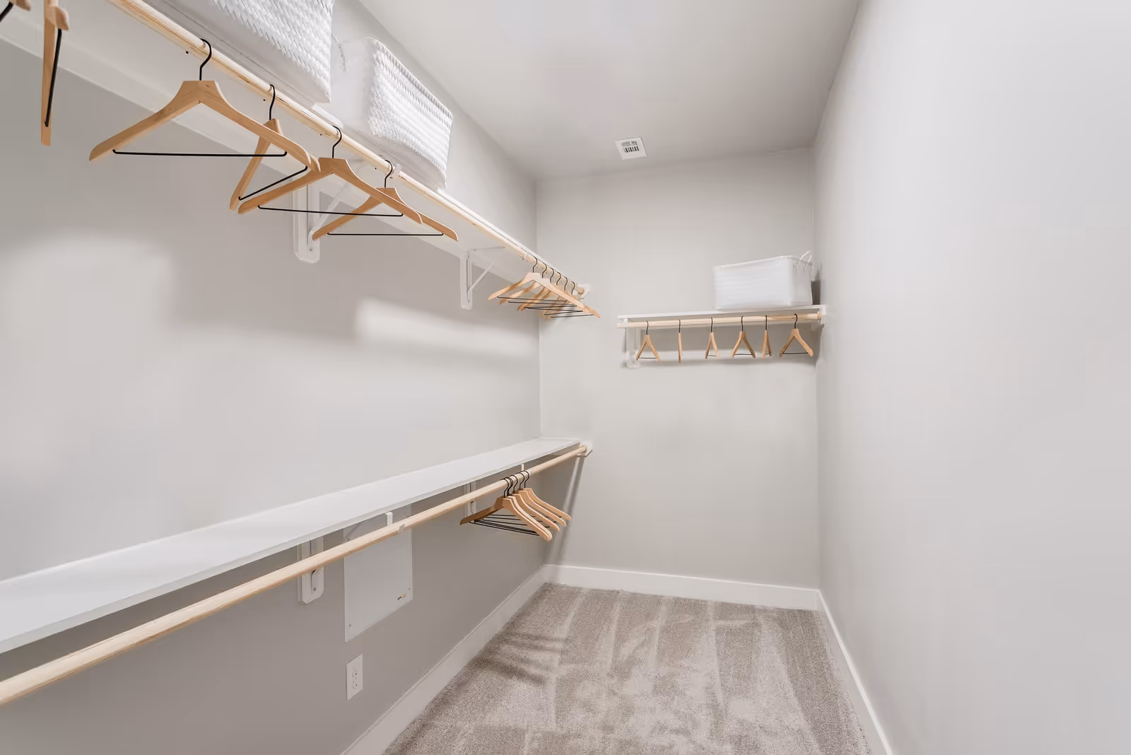 A spacious walk-in closet with light gray walls and beige carpet. The closet features two white shelves with wooden hanging rods underneath, holding several wooden hangers. There are white woven storage baskets placed on the top shelves.
