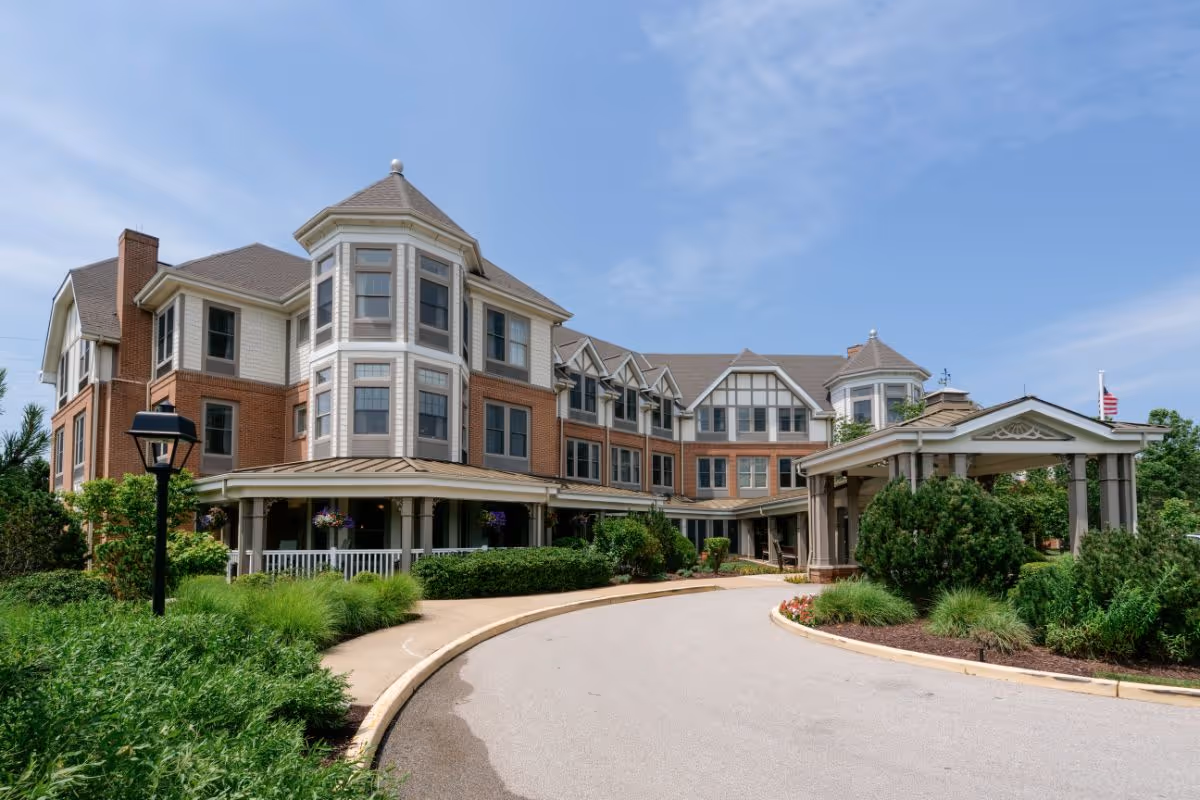 Exterior view of a large, multi-story senior living facility with a brick and beige facade, featuring multiple windows, a covered entrance with columns, and well-maintained landscaping including bushes and flowers under a partly cloudy blue sky.
