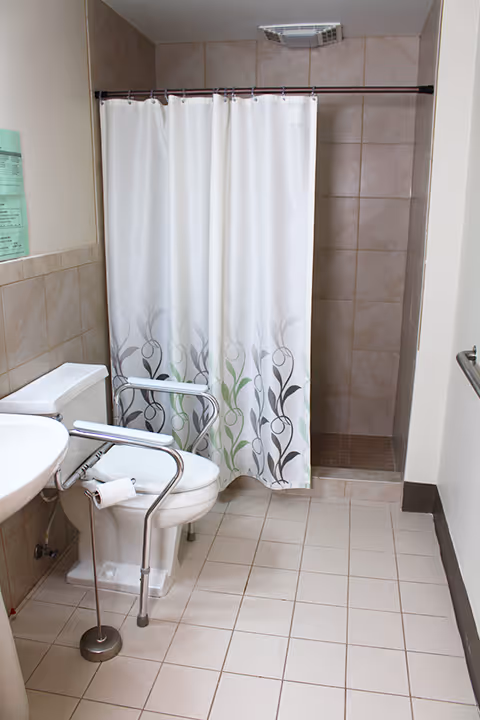 Accessible bathroom with tiled floor, a toilet fitted with safety rails, a pedestal sink, and a shower behind a white patterned curtain.