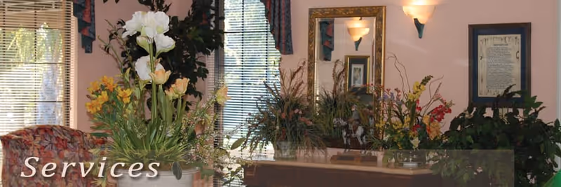 Interior view of a room with a large window covered by blinds and floral curtains, a wooden dresser with a large mirror above it, and several flower arrangements and plants placed on and around the dresser. The walls are painted light pink and decorated with framed artwork.