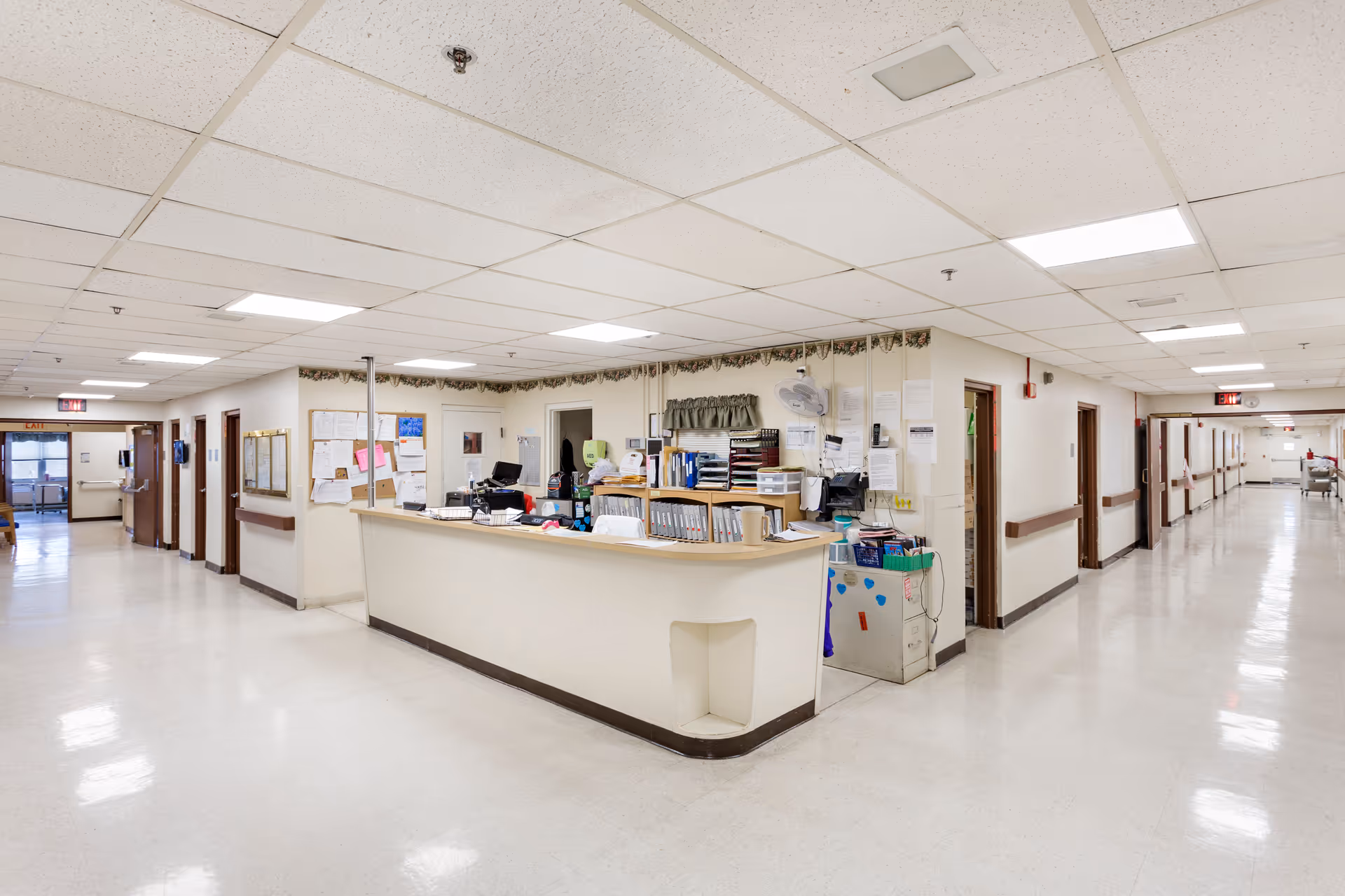 A wide, clean hallway in a senior living facility with a central nurse's station desk. The desk is cluttered with office supplies, papers, and equipment. The hallway has multiple doors leading to rooms on both sides and bright overhead fluorescent lighting.