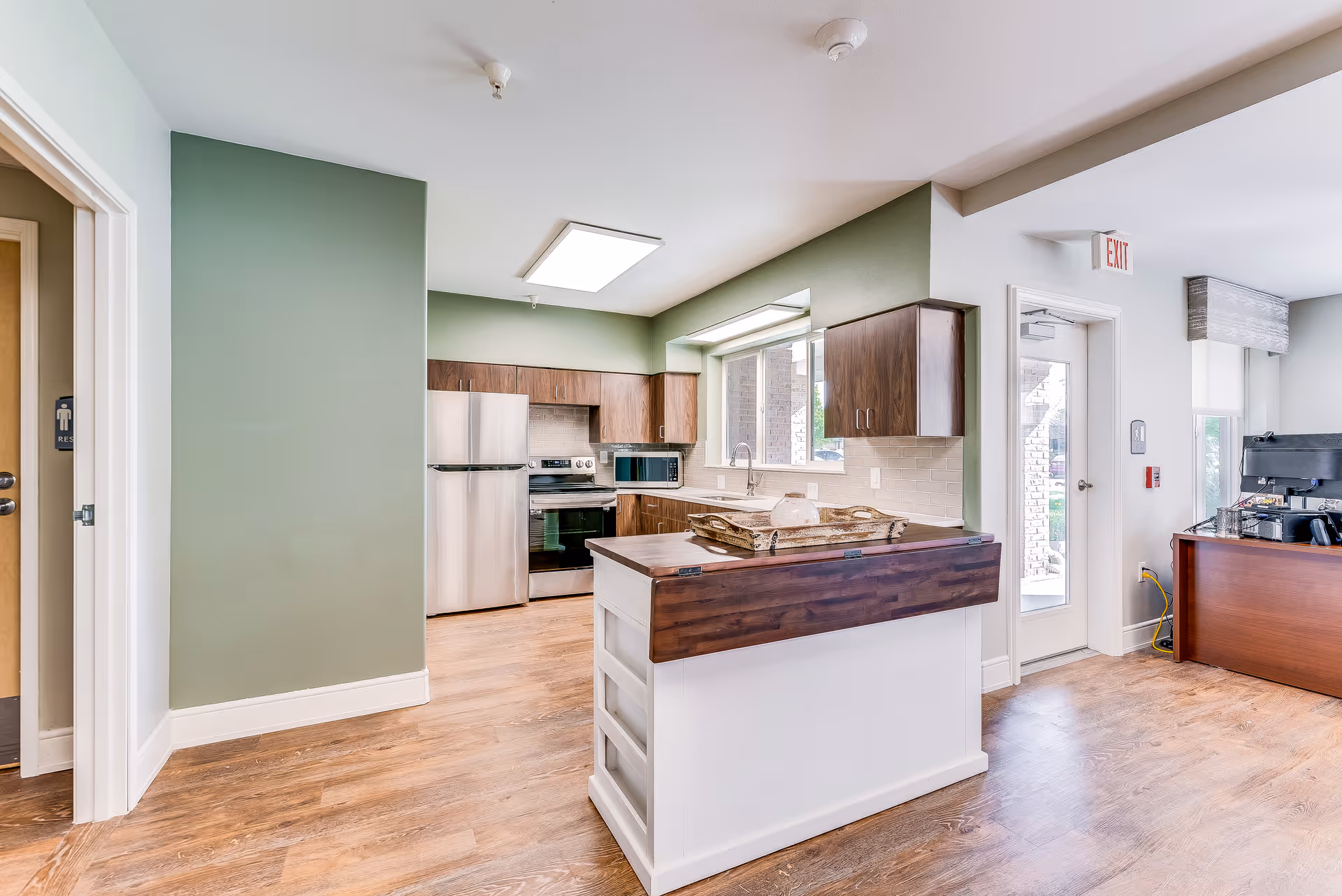 Bright and modern kitchen area in a senior living facility with wooden cabinets, stainless steel refrigerator and oven, a kitchen island with a wooden countertop, and a window letting in natural light. Adjacent to the kitchen is an office desk with computer equipment and a door leading outside.