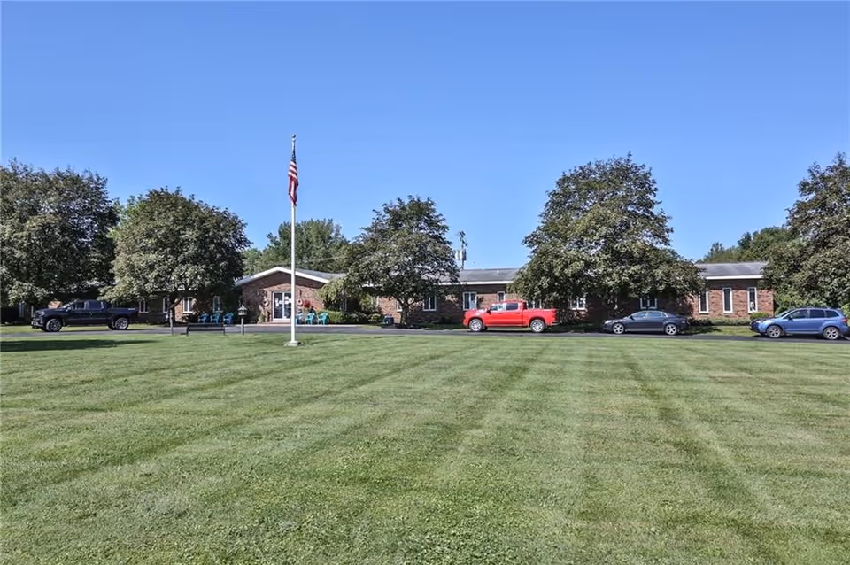 Front exterior view of Alpine Manor Home For Adults, a single-story brick building with several trees and a large green lawn in front. There is an American flag on a flagpole in the center of the lawn and several parked vehicles along the driveway in front of the building.