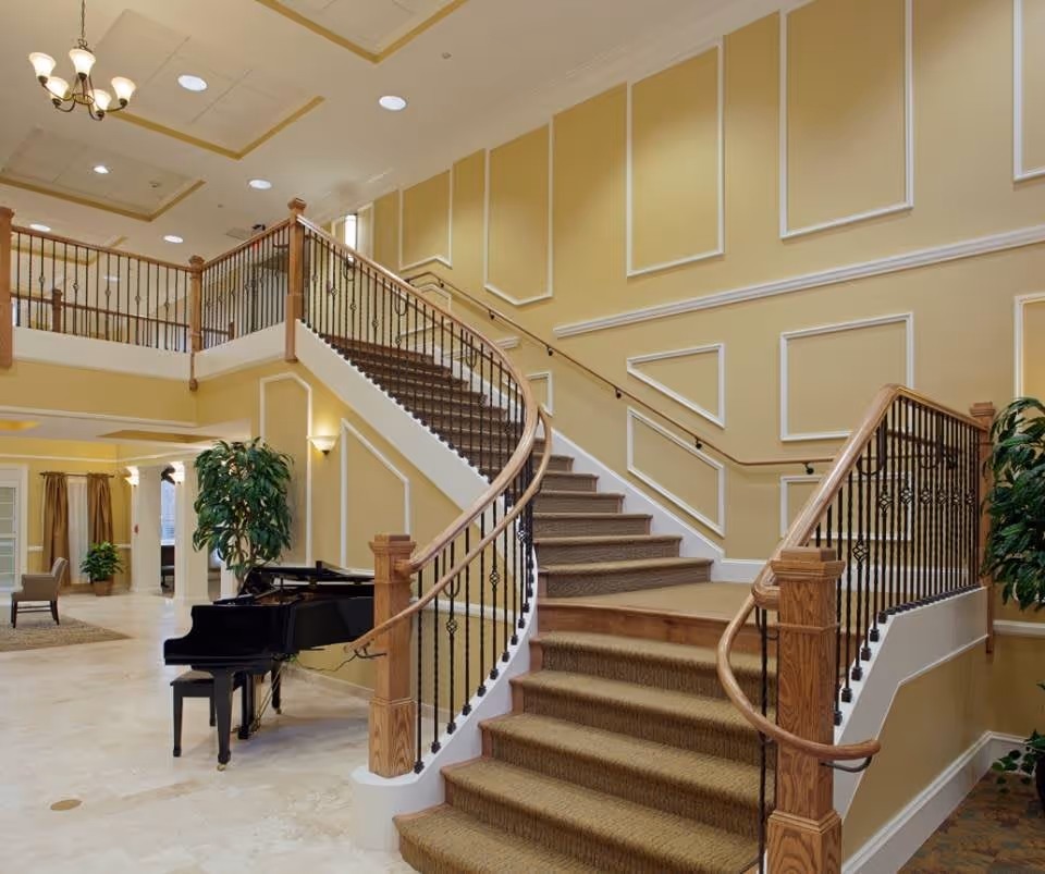 Interior view of a senior living facility featuring a grand curved staircase with wooden handrails and black iron balusters. The walls are painted yellow with white trim and decorative molding. A black grand piano is positioned near the base of the stairs, and there are green plants placed around the area. The space is well-lit with ceiling lights and a chandelier.