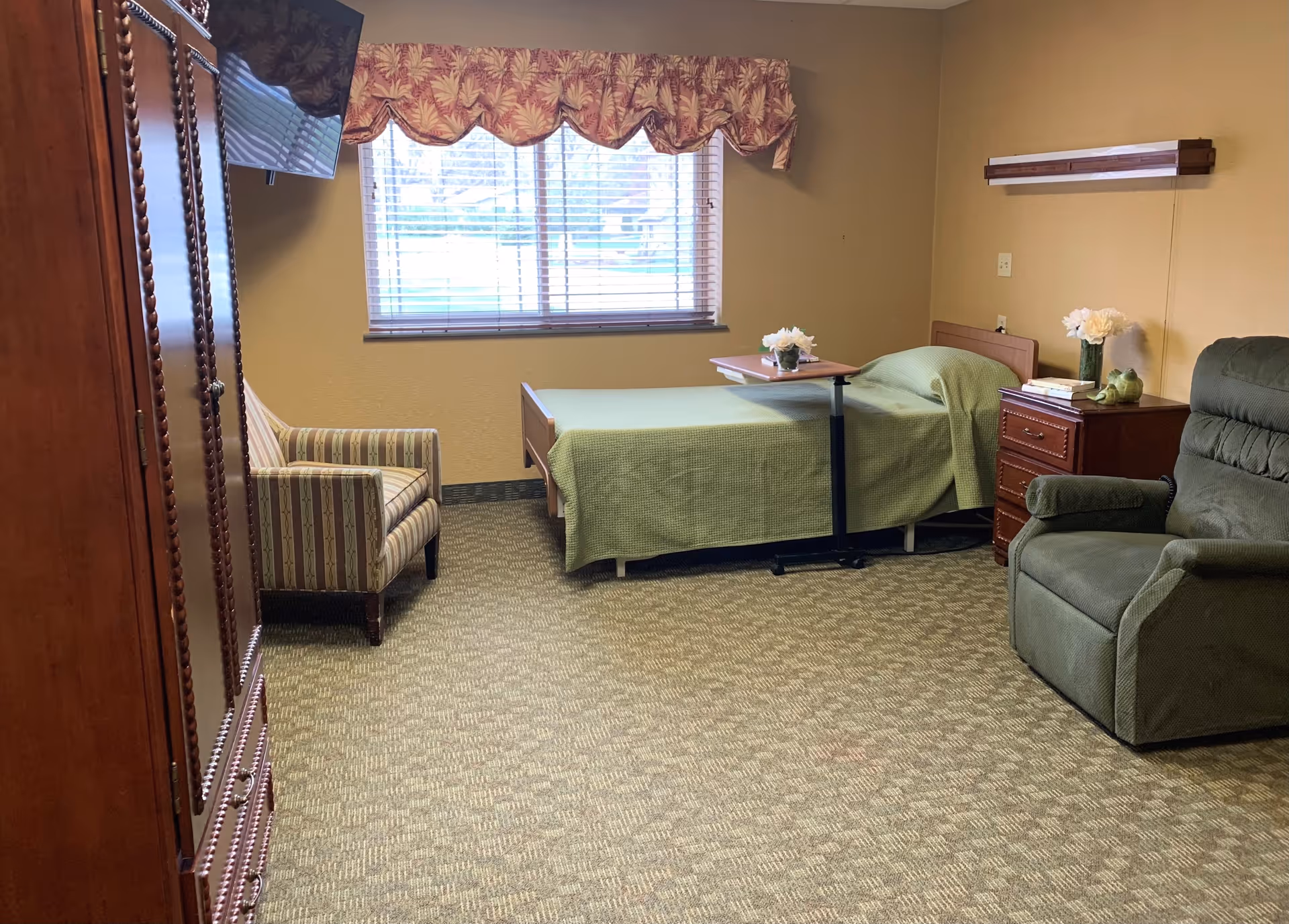 A cozy room in a rehabilitation center featuring a single bed with a green bedspread, a small overbed table with a flower vase, a wooden nightstand with books and a flower vase, a green recliner chair, a striped armchair, a wooden wardrobe, and a window with blinds and a decorative valance.