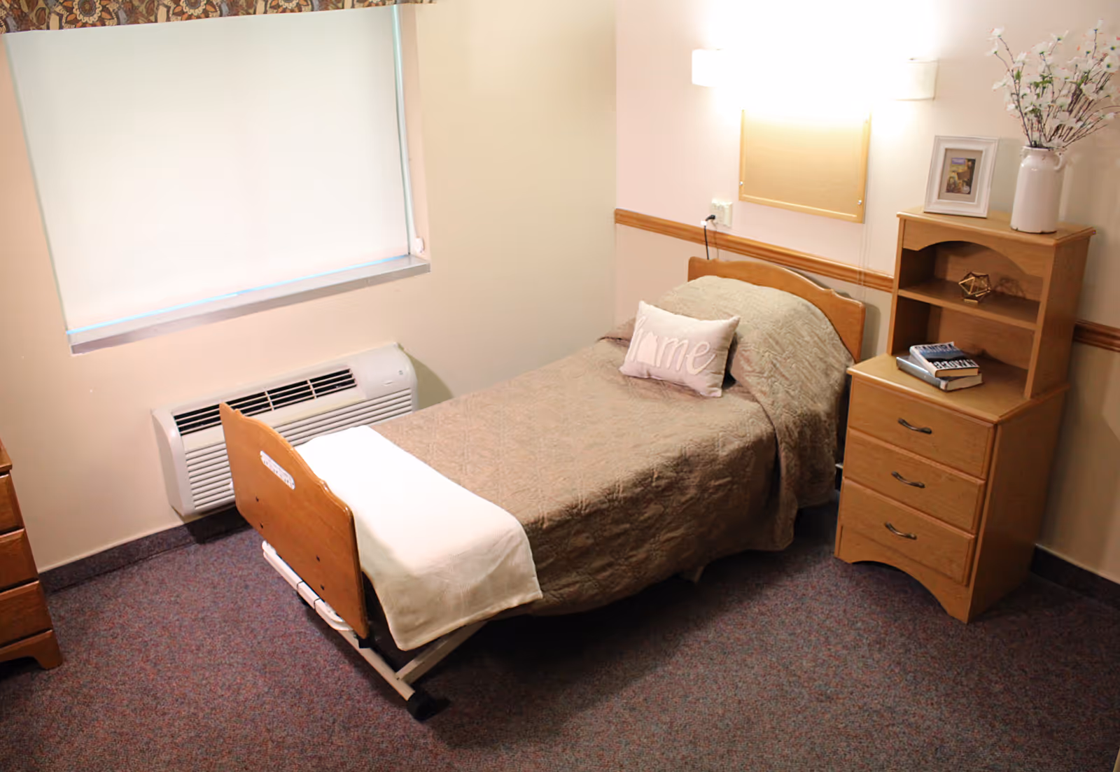A small, simple bedroom with a single bed covered in a beige quilt and a pillow with the word 'Home' on it. Next to the bed is a wooden nightstand with three drawers and an open shelf holding books, a framed photo, and a vase with flowers. A window with a closed blind and a wall-mounted heater are visible on the left side of the room.