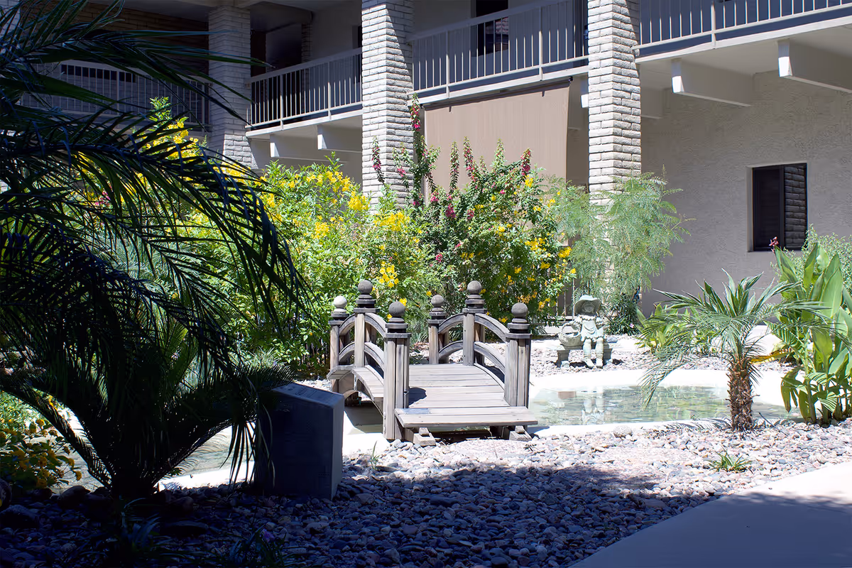 A small wooden footbridge over a shallow pond surrounded by rocks, tropical plants, and yellow flowering bushes in an outdoor courtyard area of a building with brick columns and balconies.