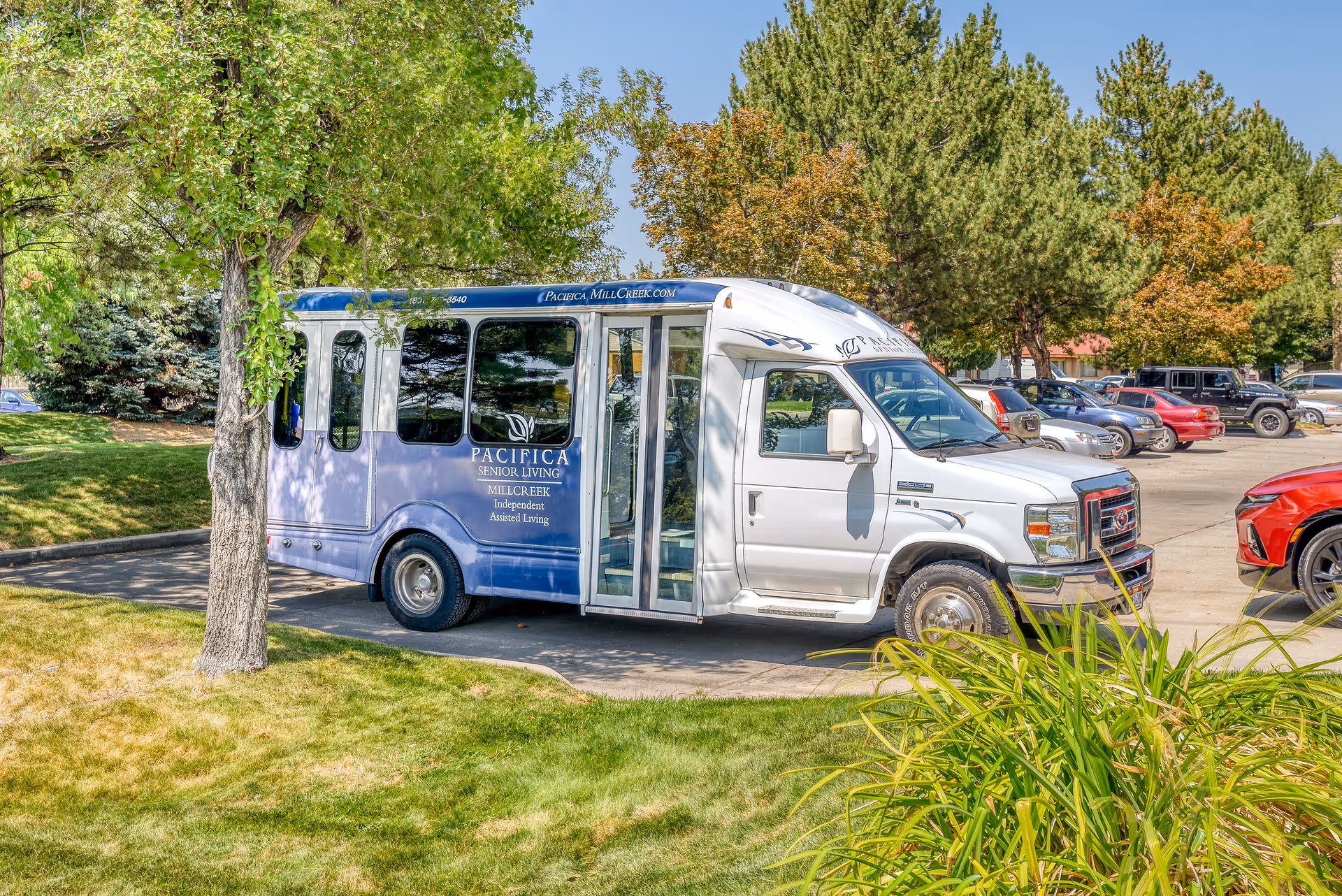 A white and blue shuttle bus parked in a parking lot surrounded by green trees and grass. The bus has signage for Pacifica Senior Living Millcreek, indicating independent and assisted living services. Several cars are parked in the background under a clear blue sky.