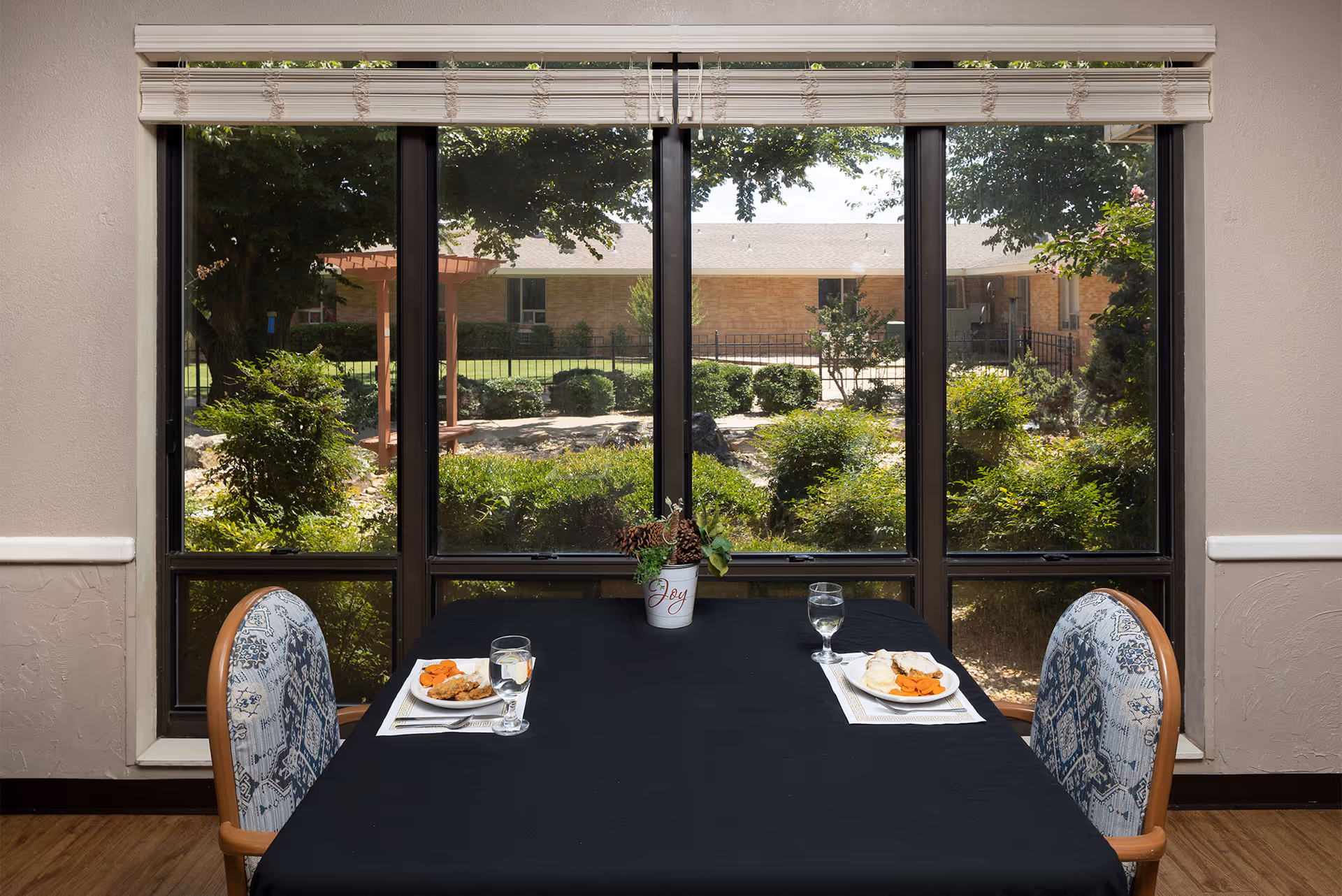 A dining table set for two with plates of food and glasses of water, positioned in front of large windows showing a garden with bushes, trees, and a pergola outside.