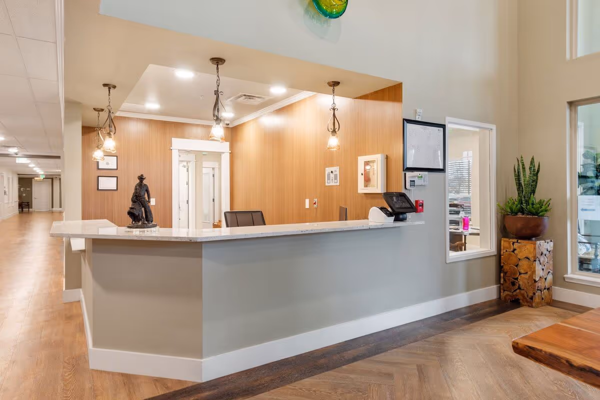 Reception desk area in a senior living facility with a light gray counter, three hanging pendant lights, a bronze sculpture on the counter, and a wooden accent wall behind. To the right, there is a window into an office and a tall plant in a wooden planter. The floor is wood with a herringbone pattern near the desk and a lighter wood tone in the hallway.