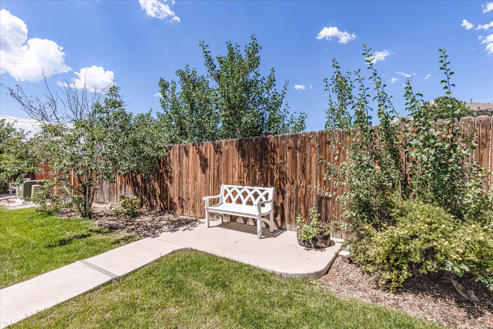 White bench on a small concrete patio beside a wooden fence in a sunny backyard with grass, trees, and shrubs.