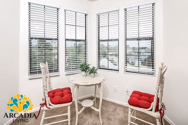 A small corner seating area with two white wooden chairs with red cushions and a small white round table between them. The area is well-lit by four tall windows with white blinds, and there are two small potted plants on the table. The floor is carpeted in a neutral tone.