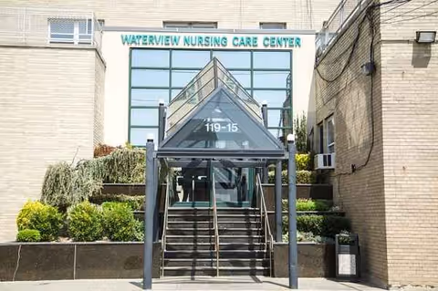 Entrance to Waterview Nursing Care Center with a glass canopy over stairs leading up to the main door, surrounded by greenery and beige brick walls.