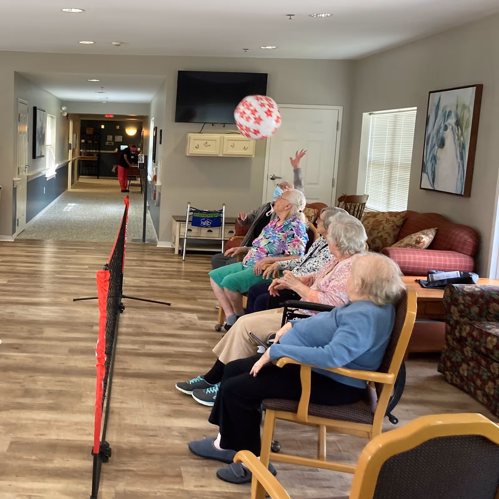 A group of elderly people sitting in chairs along a wall in a common area, playing a seated balloon volleyball game with a red and white balloon over a small net. The room has wood flooring, a TV mounted on the wall, and a couch with pillows. A staff member is visible in the hallway in the background.