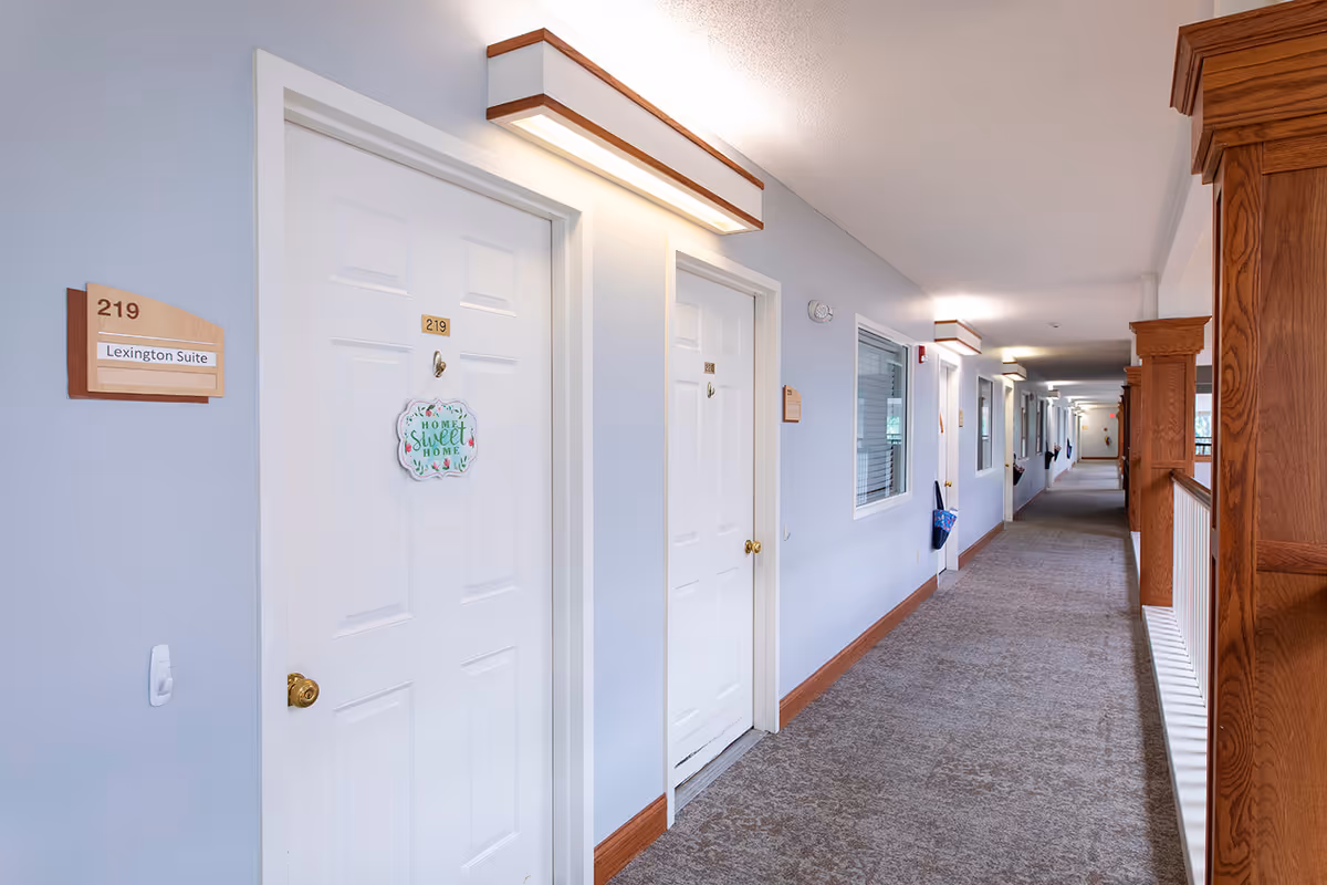Interior hallway of Lexington Park Assisted Living of Topeka with white doors numbered 219 and 220. Door 219 has a decorative sign that reads 'Home Sweet Home'. The hallway has light blue walls, carpeted floor, wooden trim, and overhead lighting fixtures. There is a window on the right wall and wooden railing along the right side.
