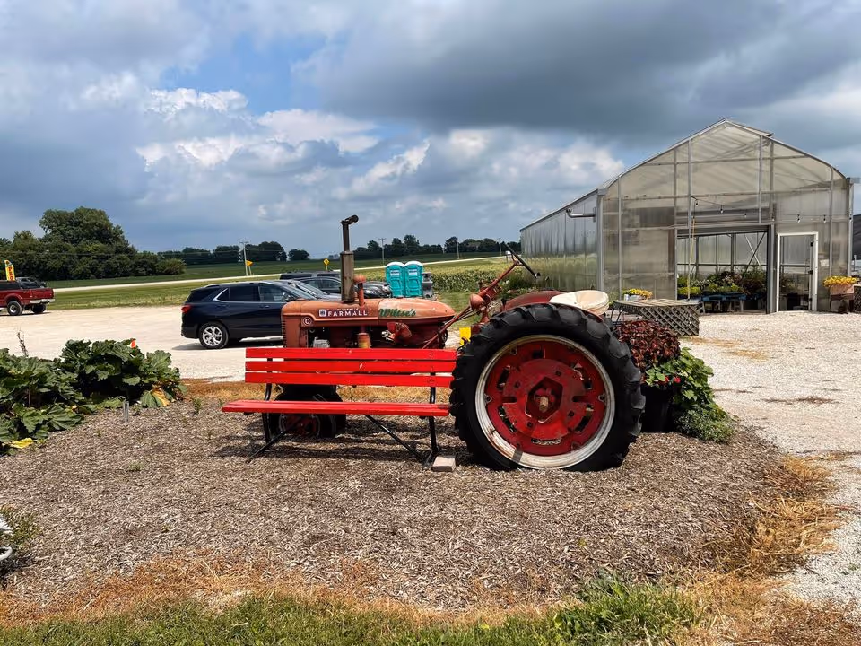 An outdoor scene featuring a vintage red Farmall tractor with a red bench attached to its side. Behind the tractor, there is a greenhouse structure and some potted plants. Several cars are parked on a gravel area, and the background shows a rural landscape with fields and trees under a partly cloudy sky.