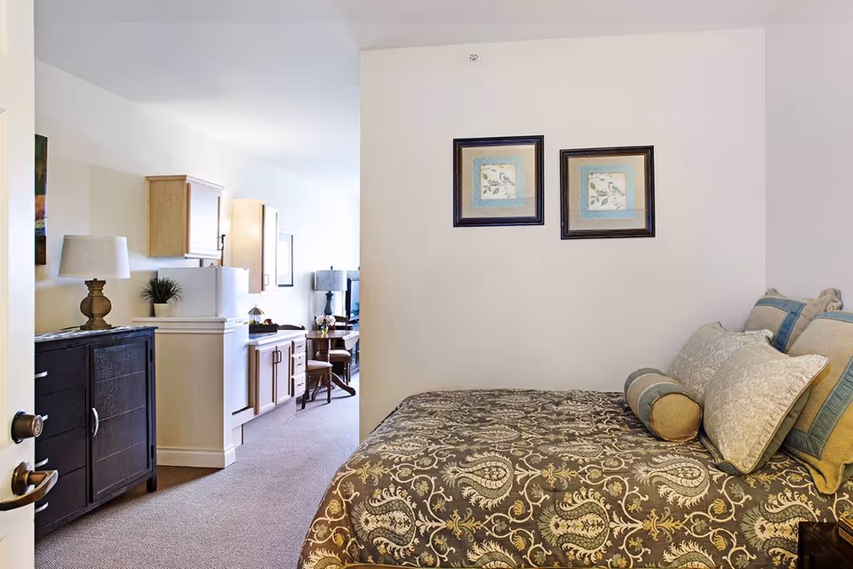 Cozy bedroom with a patterned bed and pillows, two framed pictures on the wall, and a view into a small kitchenette and sitting area.
