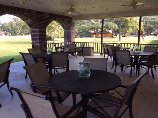 Covered outdoor patio area with multiple round tables and chairs arranged for seating. Each table has a glass jar centerpiece. The patio has brick pillars and overlooks a grassy area with trees and some buildings in the background.