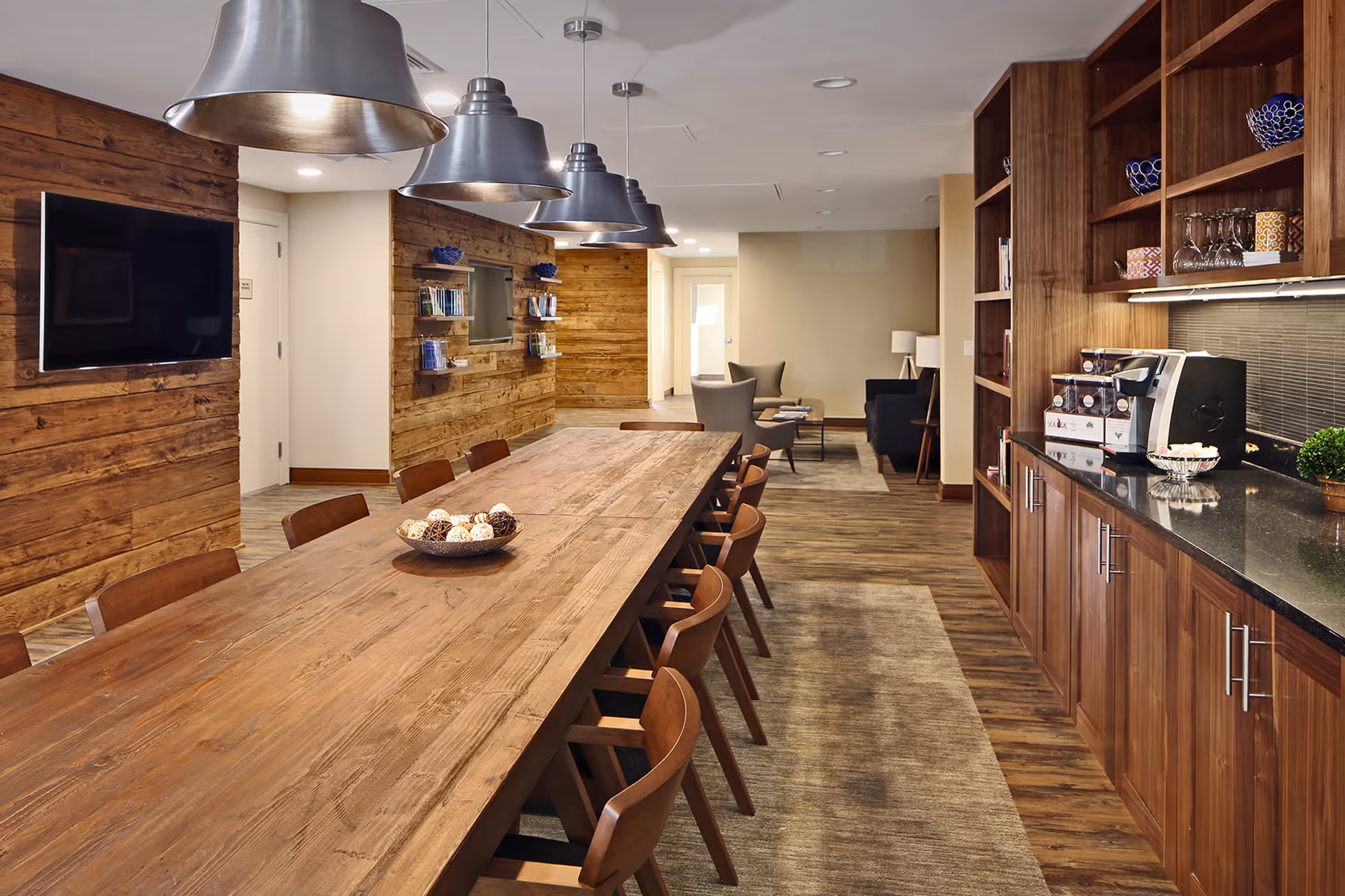 A long wooden dining table with several wooden chairs around it in a room with wooden floors and walls. Above the table are large metallic pendant lights. On the right side, there is a wooden cabinet with shelves holding decorative bowls, glasses, and a coffee machine. In the background, there is a seating area with armchairs and a lamp.