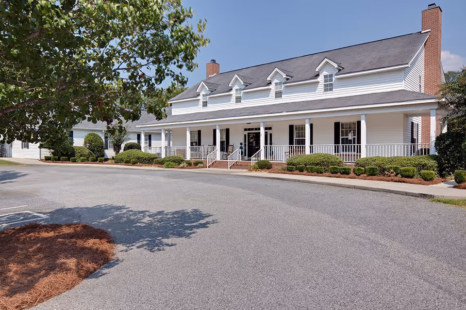 Exterior view of a large white building with a covered porch, multiple windows, and two brick chimneys. The building is surrounded by neatly trimmed bushes and trees, with a paved driveway and parking area in front under a clear blue sky.