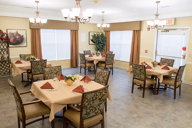 Dining room with several square tables set with beige tablecloths, folded red napkins, floral centerpieces and upholstered chairs beneath chandeliers.