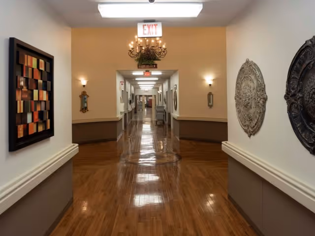 A long, polished wooden floor hallway in a healthcare facility with decorative wall art on both sides and an illuminated exit sign at the far end. The walls are painted beige with a darker trim, and there are wall sconces providing soft lighting.