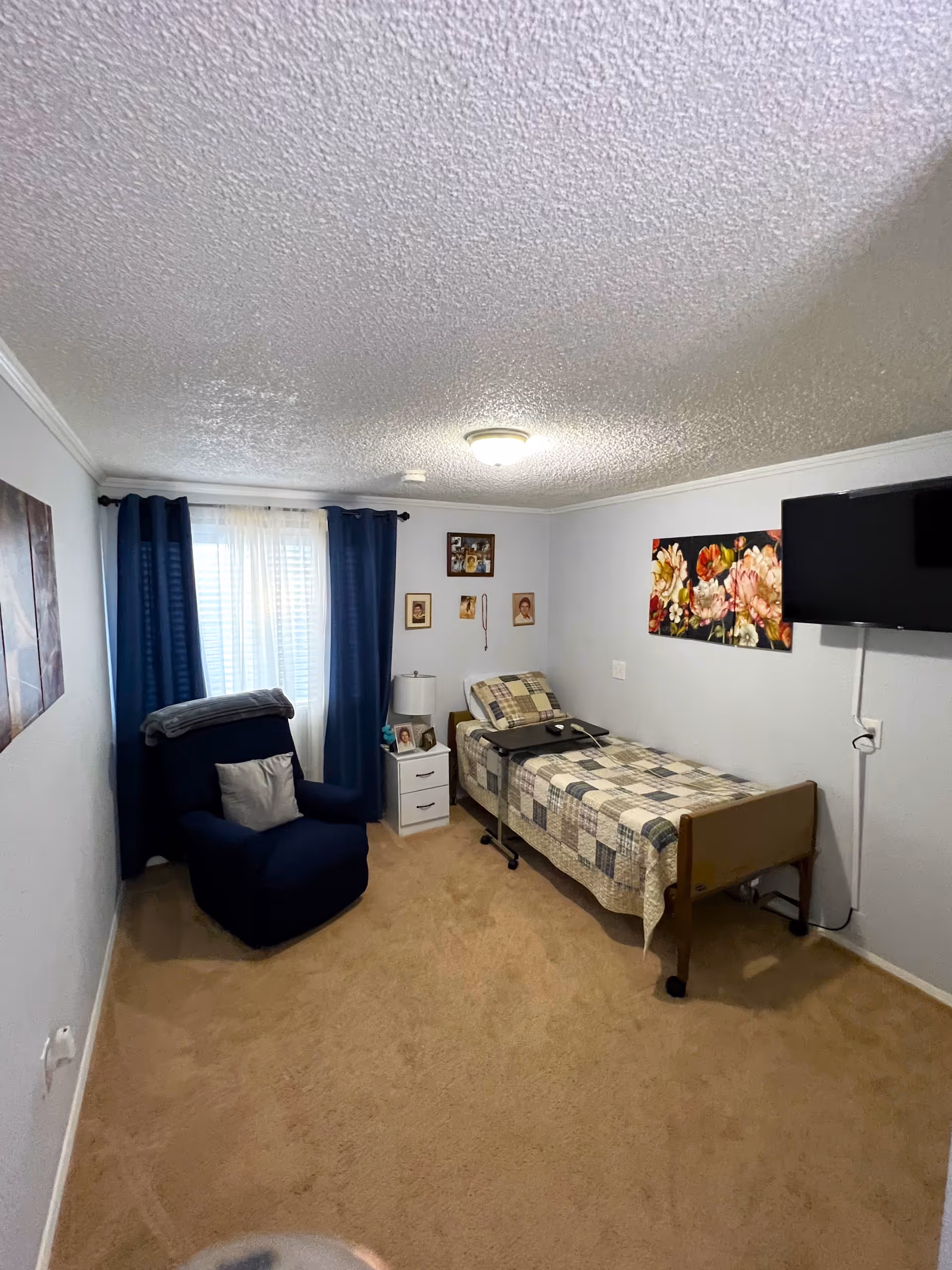 A small bedroom with beige carpet, a single bed with a patchwork quilt, a bedside table with a lamp and framed photos, a dark blue armchair with a pillow and blanket, blue curtains on a window, a wall-mounted TV, and floral artwork on the wall.