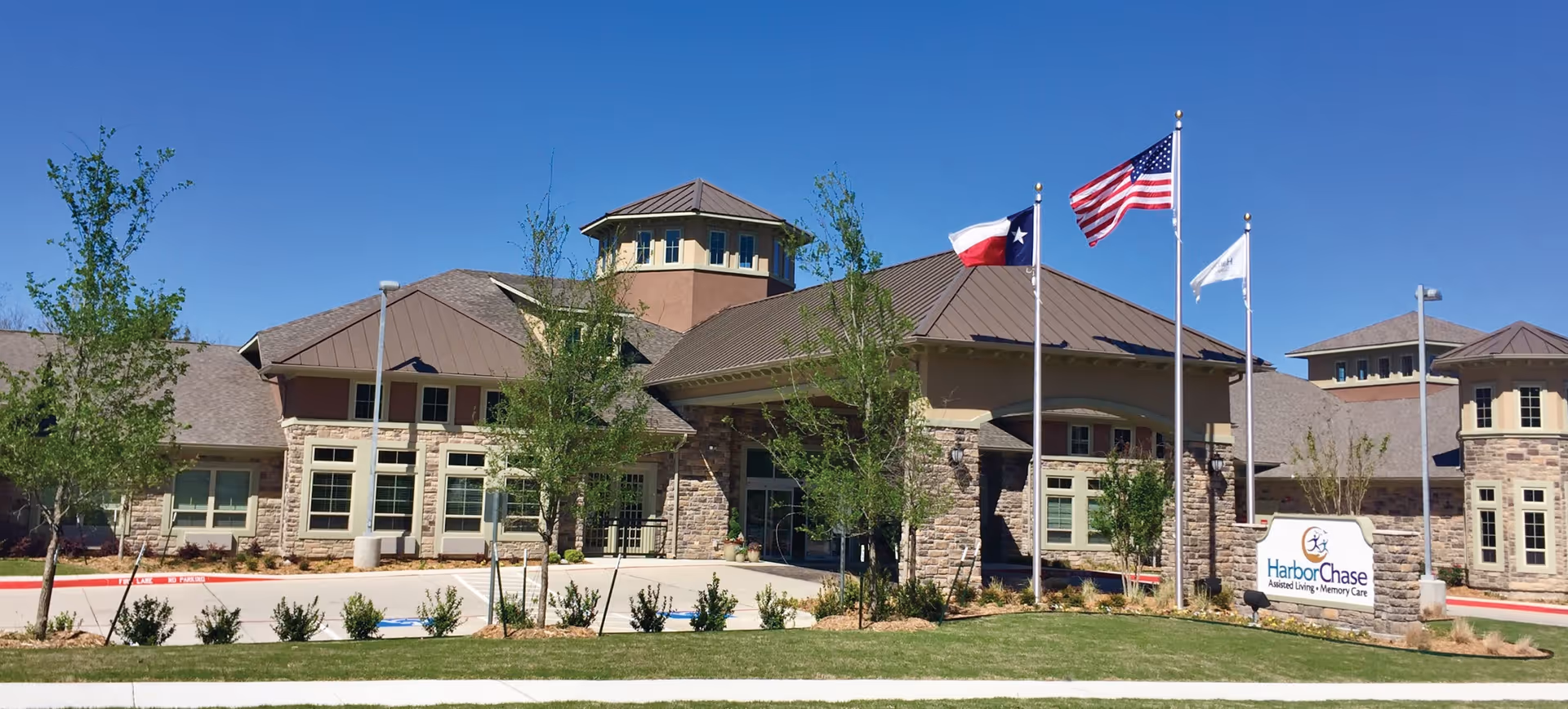 Exterior view of a senior living facility building with stone and beige walls, multiple peaked roofs, and three flagpoles displaying the Texas state flag, the American flag, and a white flag. There is a sign in front that reads HarborChase Assisted Living Memory Care. The sky is clear and blue, and there are small trees and shrubs planted around the building.