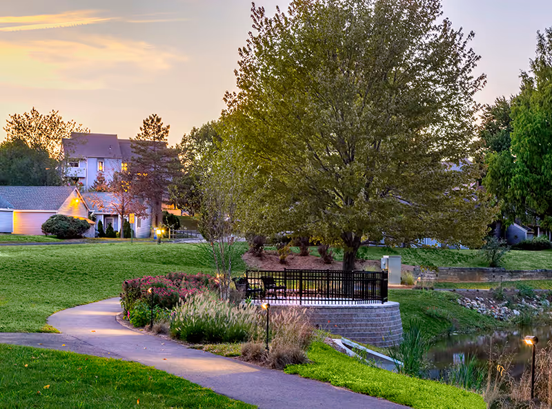 Curved paved path through landscaped lawns leading to a small pond and a railing-enclosed viewing deck beneath large trees at dusk.